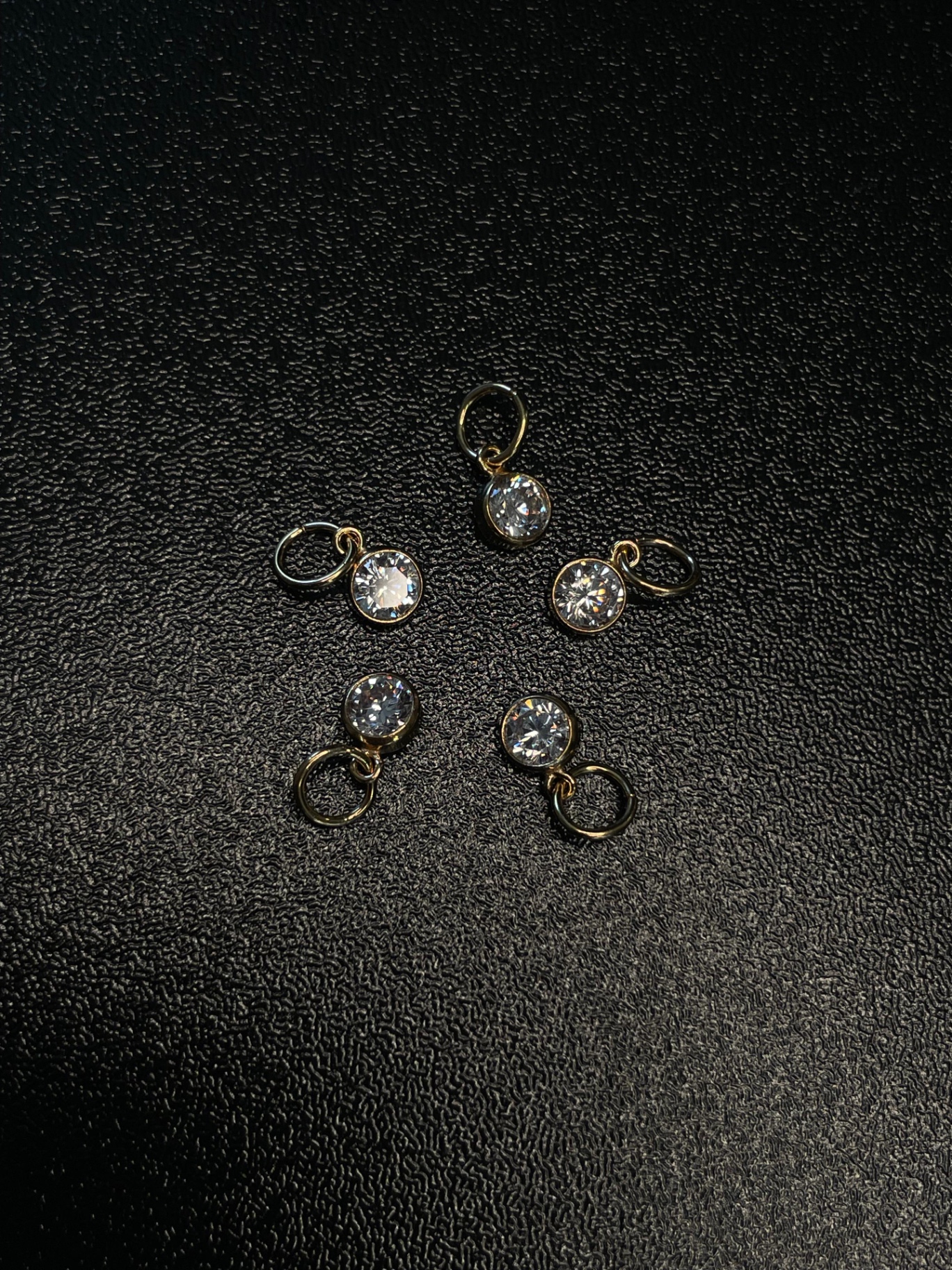 Fünf zarte Gold-Zirkonia-Charms für Permanent Jewelry auf dunklem Hintergrund